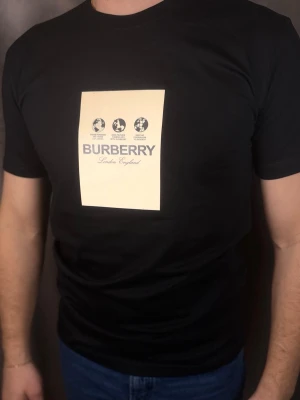 Svart Burberry t-shirt med tryck - Snygg svart t-shirt från Burberry med ett stort beige tryck på bröstet där det står 'Burberry London England' och grafiska symboler. Klassisk passform och rund hals. Perfekt för dig som gillar stilrena märkeskläder.