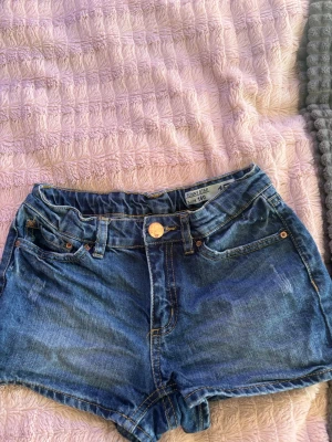 Blå denimshorts från Sunny Star - Snygga blå denimshorts från Junior Star med klassisk femficksdesign, gylf med knapp och bälteshällor. Perfekta för varma dagar och enkla att matcha med allt. Shortsen har en normal passform och är tillverkade i bomull.