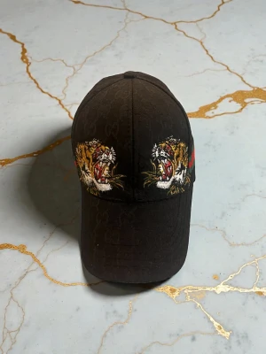 Svart Gcci keps med tigerbrodyr - Säljer en svart keps från Gcci med broderade tigrar på framsidan och sidan. Kepsen har diskret G-mönster i tyget och en klassisk böjd skärm. På sidan finns även den ikoniska gröna och röda Gucci-randen. Perfekt för dig som vill sticka ut.
