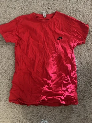 Röd Nike t-shirt med logga - En klassisk röd t-shirt från Nike med svart logga på bröstet. T-shirten har korta ärmar och rund halsringning. Perfekt för en sportig och avslappnad stil. Tillverkad i 100% mjuk bomull för skön komfort. 