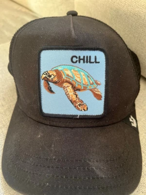 Svart keps the farm - Säljer en svart truckerkeps från Goorin Bros med mesh på sidorna och ett broderat märke framtill med en sköldpadda och texten 'CHILL' på ljusblå bakgrund. Kepsen har böjd skärm och justerbar snapback. Perfekt för en avslappnad stil.