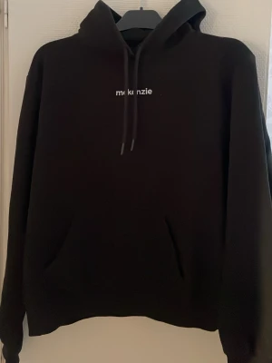 Svart hoodie från McKenzie - Säljer en svart hoodie från McKenzie med vit broderad logga på bröstet. Klassisk modell med huva och dragsnören samt känguruficka framtill. Tillverkad i mjukt material som passar perfekt för en chill och avslappnad stil.