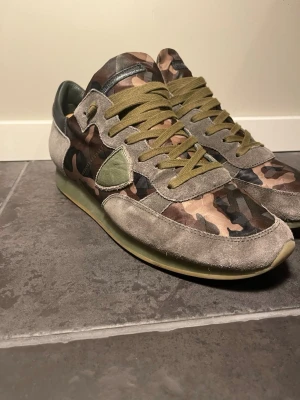 Kamouflage sneakers från Philippe Model - Snygga sneakers från Philippe Model med kamouflagemönster i grönt, brunt och svart. Ovandelen är i mocka och textil, med olivgröna skosnören och detaljer i grå mocka. Klassisk låg modell med gummisula och ikonisk sköld på sidan.