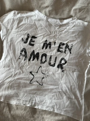 Vit t-shirt med tryck - Vit t-shirt med svart text 'JE M'EN AMOUR' och en stjärna tryckt både fram och bak. Klassisk rund hals och korta ärmar.