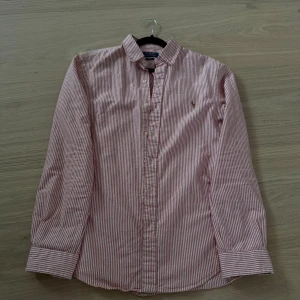 Polo Ralph Lauren skjorta - Klassisk rödvit randig skjorta från Polo Ralph Lauren i slim fit-modell. Skjortan har långa ärmar, knappar framtill och den ikoniska broderade loggan på bröstet. Tillverkad i mjuk bomull och passar perfekt för en clean och stilren look. Använd fåtal ggr eftersom att jag fick den i vintras. Priset är ej hugget i sten! Skriv vid frågor.