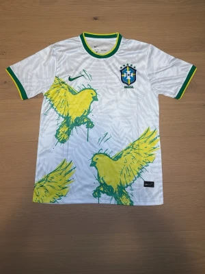 Vit Brasilien fotbollströja Nike - Snygg vit Brasilien fotbollströja från Nike med gröna och gula detaljer. Tröjan har stora gula fåglar tryckta på framsidan, gröna och gula muddar samt Brasilien-emblem på bröstet. Tillverkad i lätt och ventilerande material, perfekt för träning eller match.