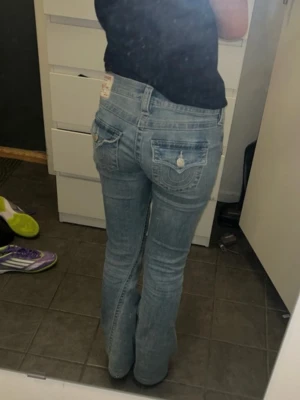 Blå bootcut jeans från True Religion - Säljer ett par ljusblå bootcut jeans från True Religion med klassiska kontrastsömmar och snygga Jeansen har låg midja och är tillverkade i mjuk denim med lätt tvättad look. De är något uppsydda så de är perfekt längd för mig som är 162 cm! Storlek: 25