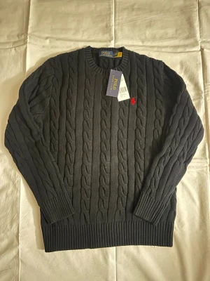 Mörkgrå kabelstickad tröja Polo Ralph Lauren - Snygg mörkgrå kabelstickad tröja från Polo Ralph Lauren med rund halsringning och klassisk röd logga broderad på bröstet. Tröjan har ribbade muddar vid ärmslut och nederkant. Perfekt för dig som gillar tidlös och stilren design.