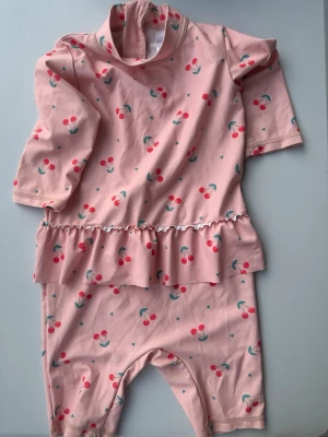 Rosa baddräkt med körsbärsmönster86/92 - Supersöt rosa baddräkt för barn med körsbärsmönster och volang i midjan. Dräkten har långa ärmar och ben, samt dragkedja i ryggen för enkel påklädning. Materialet är stretchigt och bekvämt, perfekt för lek i vattnet.