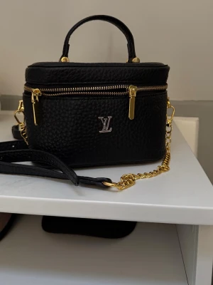 Svart handväska - Snygg svart handväska   i strukturerat läder med guldfärgade detaljer och kedja. Väskan har ett topphandtag, avtagbar axelrem och dragkedja. Insidan är fodrad i beige tyg och har ett fack. Perfekt för dig som gillar lyxiga accessoarer.