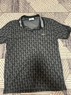 Svart mönstrad piké från Lacoste - Svart pikétröja från Lacoste med grått geometriskt mönster och klassisk krage med vita ränder. Ikoniska gröna krokodilloggan på bröstet. Skön passform och perfekt för en stilren look.