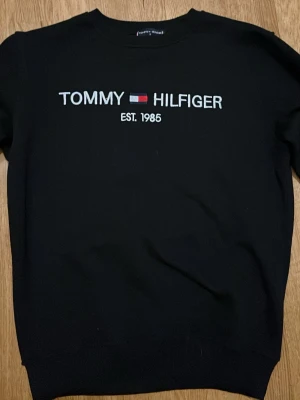 Svart sweatshirt Tommy Hilfiger - Svart sweatshirt från Tommy Hilfiger med broderad logga och text 'EST. 1985' på bröstet. Klassisk rund halsringning och ribbade muddar vid ärmslut och nederkant. Liten flaggdetalj på ärmen. Perfekt för en clean och stilren look.