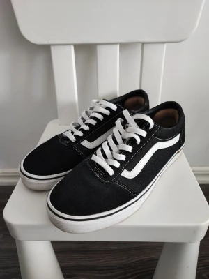 Svarta Vans Old Skool sneakers - Klassiska Vans Old Skool sneakers i svart canvas och mocka med vit sula och vit sidostripes. Skorna har vita snören och låg profil som ger en clean och tidlös look. Perfekta för dig som gillar streetwear och skate-inspirerad stil.