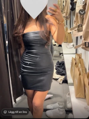 Svart axelbandslös kort klänning - Säljer en svart, tight och axelbandslös kort klänning i ett glansigt material. Klänningen har en kroppsnära passform och är perfekt för dig som vill ha en stilren och trendig look.