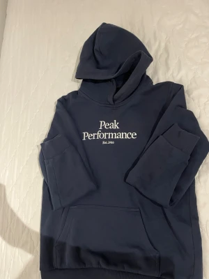 Mörkblå hoodie från Peak Performance - Snygg mörkblå hoodie från Peak Performance med vit broderad logga på bröstet. Klassisk känguruficka framtill och stor huva. Tillverkad i mjukt bomullsmaterial som är skönt att ha på sig. Perfekt för en avslappnad och sportig stil.