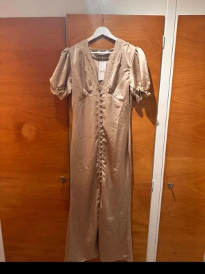 Champagnefärgad långklänning  - Elegant långklänning i champagnefärg från Byic med v-ringning, spetsdetaljer och knappar framtill. Klänningen har korta puffärmar och är tillverkad i ett glansigt material som ger en lyxig känsla. Perfekt för speciella tillfällen.