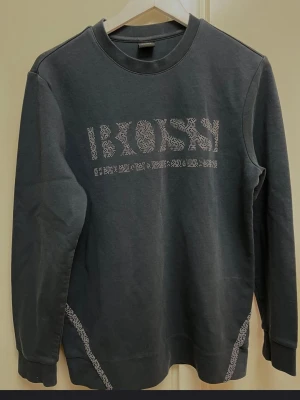 Mörkgrå sweatshirt från BOSS - Snygg mörkgrå sweatshirt från BOSS med vit logotyp i mönstrade bokstäver på bröstet. Tröjan har rund halsringning, långa ärmar och dekorativa vita detaljer vid sidorna. Perfekt för en avslappnad och trendig stil.