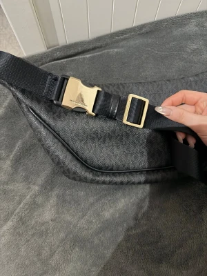 Svart axelväska från Michael Kors - Snygg svart axelväska från Michael Kors med guldiga detaljer och dubbla dragkedjor. Väskan har justerbart band med spänne och är tillverkad i ett tåligt material med MK-logga över hela ytan. Perfekt för att bära det viktigaste med stil.