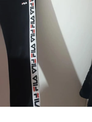 Svarta mjukisbyxor från FILA - Säljer ett par svarta mjukisbyxor från FILA med vita sidor där FILA-loggan upprepas i rött, blått och svart. Byxorna har resår i midjan och raka ben. Perfekta för chill eller träning och har en sportig vibe.