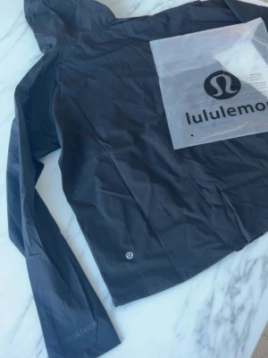 Svart träningsjacka från Lululemon - Snygg svart träningsjacka från Lululemon med huva och dragkedja framtill. Tillverkad i ett lätt och stretchigt material som passar perfekt för löpning eller gym. Diskret logga på bröstet och ärmen samt praktiska fickor med dragkedja.
