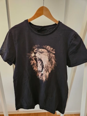 Svart t-shirt med lejontryck - Cool svart t-shirt med ett stort tryck av ett rytande lejon på bröstet. Klassisk rund halsringning och korta ärmar. Perfekt för dig som vill sticka ut med ett djurmotiv. Tillverkad i mjuk bomull för skön känsla hela dagen.