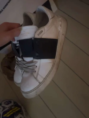 Valentino vita sneakers med svart band - Säljer ett par snygga vita sneakers från Valentino med ett brett svart band över sidan och klassisk snörning. Skorna har en chunky sula och är tillverkade i skinn för en lyxig känsla. Perfekta för dig som vill ha en stilren och trendig look.
