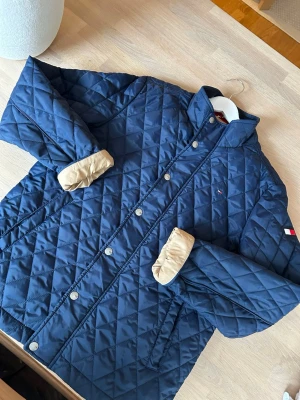 Marinblå quiltad jacka Tommy Hilfiger - Snygg marinblå quiltad jacka från Tommy Hilfiger med tryckknappar och ståkrage. Jackan har diskret logga på bröstet och flagga på ärmen. Fodret är beige vid ärmsluten och materialet är mjukt och vadderat för extra komfort.