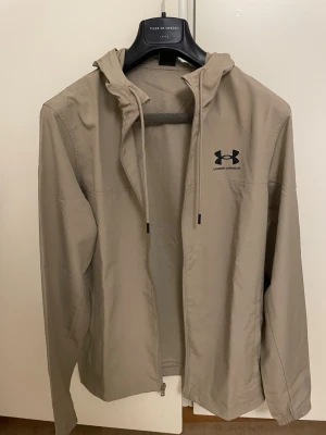 Beige vindjacka från Under Armour - Stilren beige vindjacka från Under Armour med dragkedja framtill och justerbar huva med snören. Jackan har svart logga på bröstet och är tillverkad i ett lätt, vindtätt material som passar perfekt för blåsiga dagar.