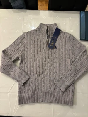 Ljusgrå kabelstickad tröja Polo Ralph Lauren - Ljusgrå kabelstickad tröja från Polo Ralph Lauren med halv dragkedja och hög krage. Tröjan har ribbade muddar och nederkant samt den klassiska broderade loggan på bröstet. Perfekt för dig som gillar stilren och tidlös design.