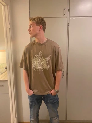 Beige  Ance studios t-shirt - Säljer en beige t-shirt från Acne studios med ett självreflekterande tryck på bröstet. T-shirten har en loose passform och är tillverkad i mjuk bomull. Perfekt för en avslappnad och trendig stil.