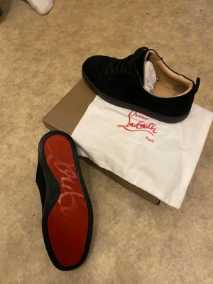 Svarta Christian Louboutin sneakers - Snygga svarta sneakers från Christian Louboutin i mocka med klassisk röd sula och svarta skosnören. Insidan är beige och skon har en stilren, låg profil som passar till många olika outfits. Ikonisk logga på sulan och exklusiv känsla.