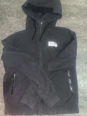 Svart vindjacka från EA7 Emporio Armani - Snygg svart vindjacka från EA7 Emporio Armani med huva, hel dragkedja framtill och två fickor med dragkedja. Jackan har en stilren logga på bröstet och justerbara snören i huvan. Perfekt för dig som vill ha en sportig och clean look. Köpt för 1600kr.