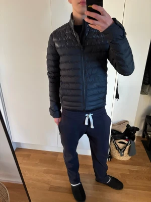 Svart dunjacka från J.Lindeberg - Snygg svart dunjacka från J.Lindeberg med quiltad design och hög krage. Jackan har dragkedja framtill och två sidofickor. Perfekt för kalla vinterdagar och har en stilren, smal passform. Materialet är lätt och värmande dun.