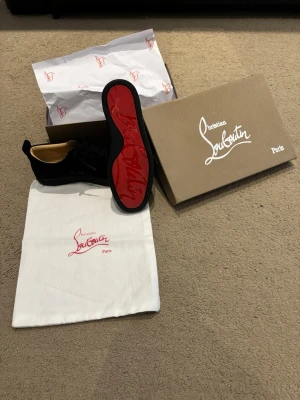 Svarta Christian Louboutin - Snygga svarta sneakers från Christian Louboutin med klassisk röd sula och svarta skosnören. Ovandelen är i mocka och insidan är beige. Kommer med originalkartong och dustbag. Perfekta för dig som vill ha något exklusivt och stilrent.