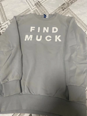 Grå sweatshirt från H&M Divided - Säljer en grå sweatshirt från H&M Divided med texten 'FIND MUCK' i vitt på bröstet. Tröjan har rund hals, ribbade muddar vid ärmslut och nederkant samt en avslappnad passform. Perfekt för en chill och casual stil. Lite fläck men tror går o tvättas bort