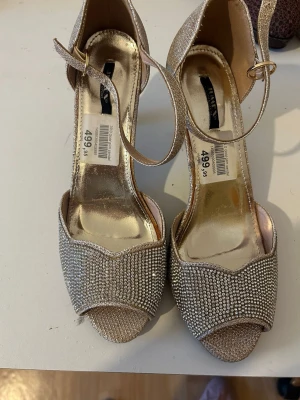 Glittriga pumps med rem från NLY Shoes - Säljer ett par glittriga pumps från NLY Shoes i champagnefärg med öppen tå och smal ankelrem. Skorna är täckta av små strassdetaljer framtill och har en elegant klack. Perfekta för att sticka ut och ge extra bling till din outfit.