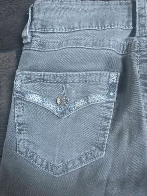 Grå jeans med dekorerad bakficka - Snygga grå jeans med raka ben och unik dekorerad bakficka med silverfärgad knapp och mönstrad kant. Jeansen har klassisk femficksdesign och är tillverkade i mjukt denimtyg. Perfekta för dig som vill ha något extra i din jeansgarderob.