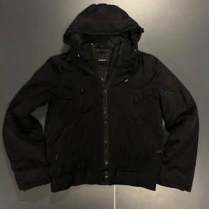 Svart jacka med huva från Fishbone - Svart jacka från Fishbone med huva och dragkedja framtill. Jackan har flera fickor, dragsko i huvan och ribbade muddar vid ärmslut och midja. Perfekt för kyliga dagar och har en sportig, avslappnad stil. Storlek M i tjej men passar S-XS i man beroende på hur man vill att den sitter.