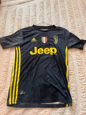 Juventus Ronaldo matchtröja svart/gul - Juventus matchtröja från Adidas i svart med gula detaljer och tryck. Framsidan har klubbmärke, Adidas-logga och Jeep som sponsor. Baksidan har Ronaldo och nummer 7 i gult. Tillverkad i lätt och ventilerande material, perfekt för fotbollsträning eller match.