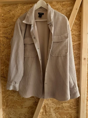 Beige overshirt i manchester från Lindex - Stilren beige overshirt från Lindex i mjuk manchester. Modellen har två stora bröstfickor, knappstängning framtill och avslappnad passform. Perfekt lager-på-lager-plagg med trendig oversized look och bred krage.