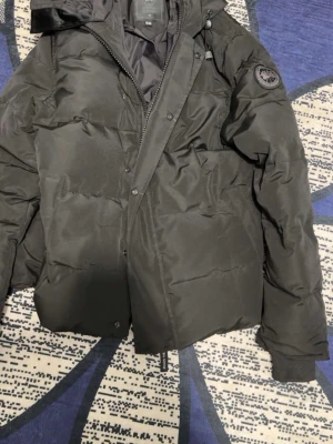 Svart pufferjacka från Canada Goose - Säljer en svart pufferjacka från Canada Goose med huva och ribbade muddar. Jackan har en klassisk patch-logga på ärmen och dragkedja med knappslå. Perfekt för kalla vinterdagar och riktigt varm tack vare det tjocka materialet. Size XL skriv din längd 