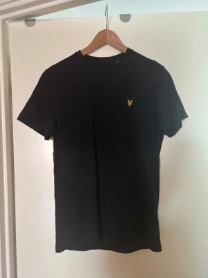 Svart t-shirt från Lyle & Scott - Klassisk svart t-shirt från Lyle & Scott med gul broderad logga på bröstet. T-shirten har rund halsringning och korta ärmar. Tillverkad i mjuk bomull för en bekväm känsla och passar perfekt till alla typer av outfits.