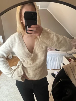 Beige omlottstickad tröja - Mysig beige stickad tröja med omlottmodell och djup v-ringning. Tröjan har långa ärmar och ribbstickade muddar vid ärmslut och nederkant. Perfekt för lager-på-lager och enkel att styla till olika outfits.