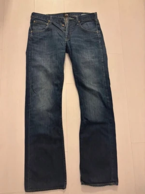 Lee blå raka jeans W31 L34 - Säljer ett par klassiska blå jeans från Lee i modellen Flint. Jeansen har raka ben, fem fickor och gylf med knapp och dragkedja. Tillverkade i slitstarkt denimtyg, perfekta för en avslappnad och tidlös stil.