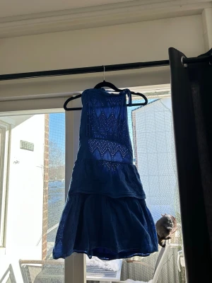 Blå halternäck klänning med spetsdetaljer - Säljer en blå kort klänning med spetsdetaljer och volanger. Klänningen har knyt i halsen och öppen rygg, samt ett vackert broderat mönster. 