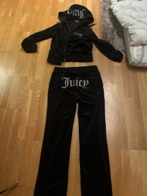 Juicy Couture svart velourset - Juicy Couture loungewear set i svart velour med dragkedja på hoodien och fickor framtill. Setet har glittrande Juicy-tryck i strass på både huvan, bröstet och byxans bakficka. Perfekt för en avslappnad och trendig look.