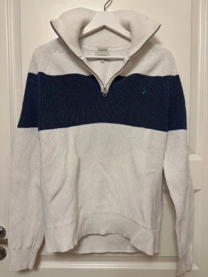 Halvzip tröja från Boomerang - Stickad tröja från Boomerang i vitt med bred marinblå rand över bröstet. Halvzip-dragkedja och hög krage ger en sportig look. Liten logga broderad på bröstet. Ribbstickade muddar ärmarna och nederkant. Perfekt för lager på lager.