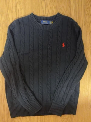 Svart stickad polo Ralph lauren tröja - Kabelstickad svart tröja från Polo Ralph Lauren med rund halsringning och röd broderad logga på bröstet. Tröjan har ribbade muddar vid ärmslut och nederkant och är tillverkad i mjukt material. Perfekt för dig som gillar klassisk och stilren look.