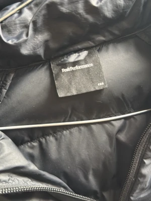 Svart pufferjacka från peak performance  - Snygg svart pufferjacka med huva och dragkedja framtill. Jackan har quiltad design, breda muddar vid ärmslut och nederkant samt logga på bröstet. Perfekt för kalla vinterdagar och riktigt skön att bära.