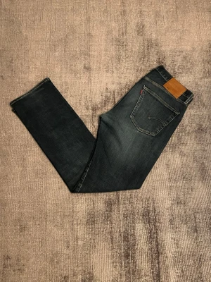 Mörkblå Levi's jeans - Säljer ett par mörkblå jeans från Levi's med klassisk femficksmodell och en snygg slitning på ena benet. Jeansen har raka ben och en brun läderpatch bak i midjan. Perfekta för en avslappnad och trendig look.